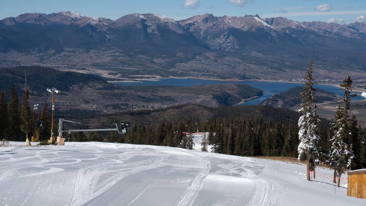 Snow-covered ski slope with pine trees and distant mountains under blue sky.