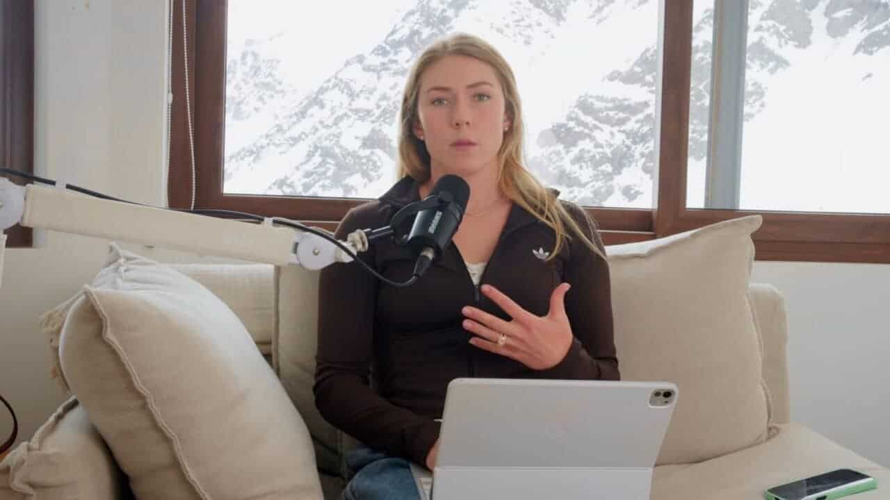 Woman speaking into microphone with laptop and snow-covered window behind her.