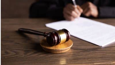 Gavel resting on a wooden desk with a person writing in the background.