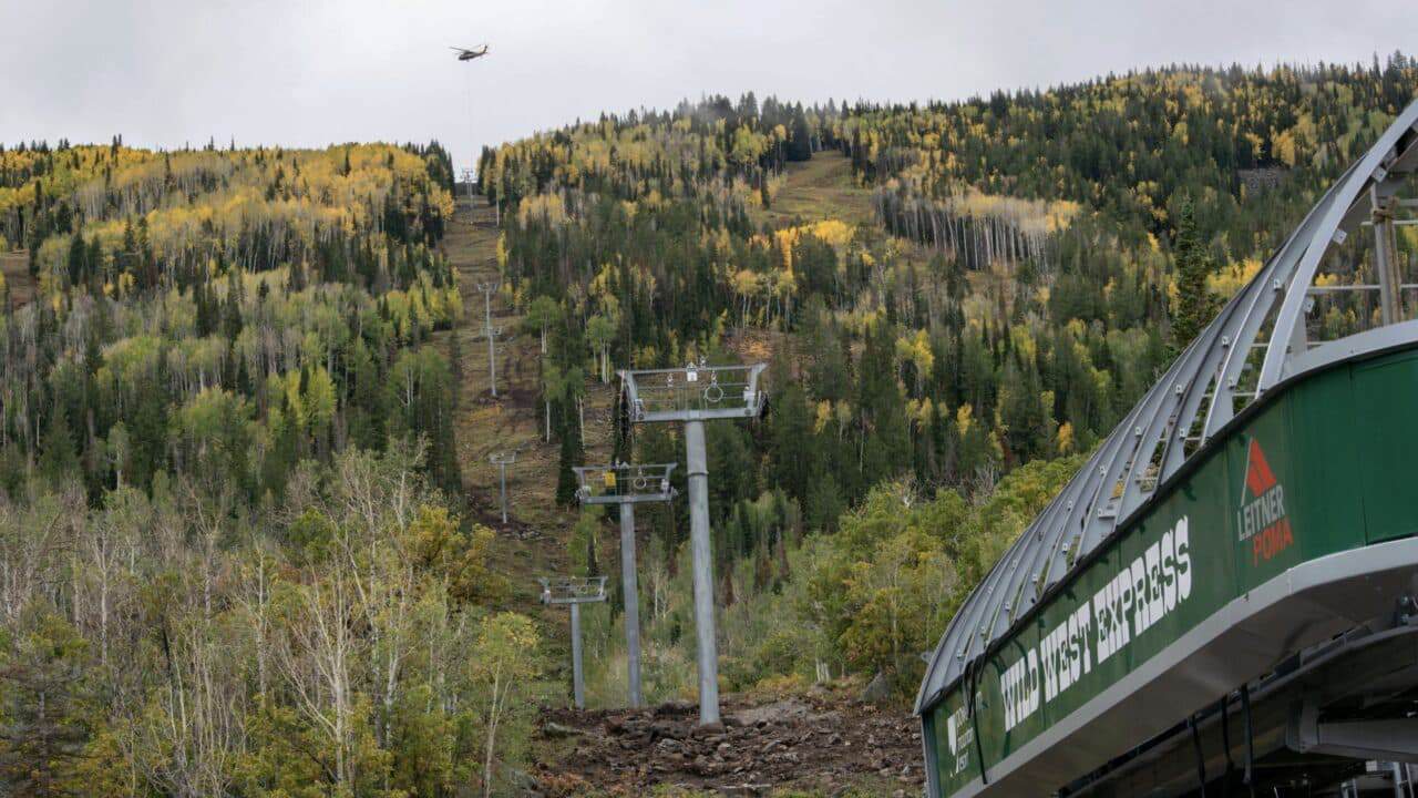 Ski lift ascending through colorful autumn forest with helicopter overhead.