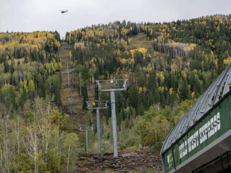 Ski lift ascending through colorful autumn forest with helicopter overhead.