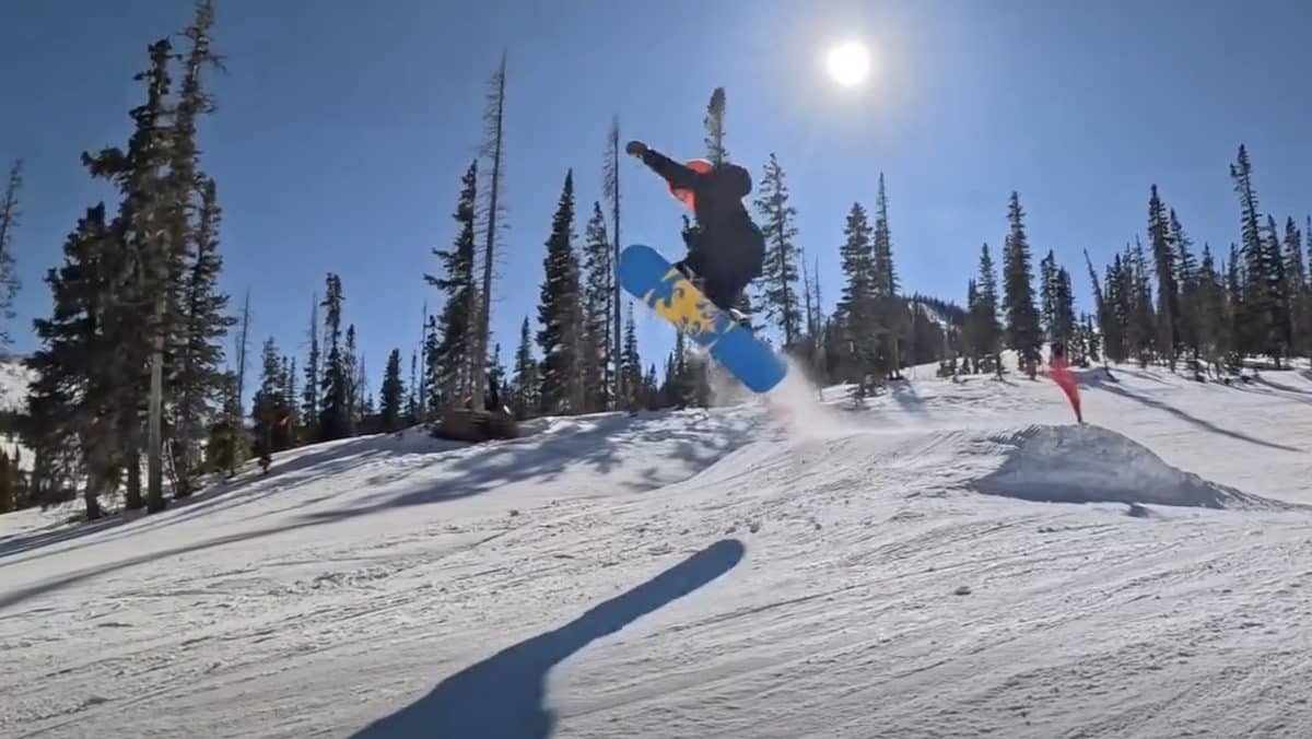 Snowboarder mid-air jump on snowy mountain with pine trees and sun.