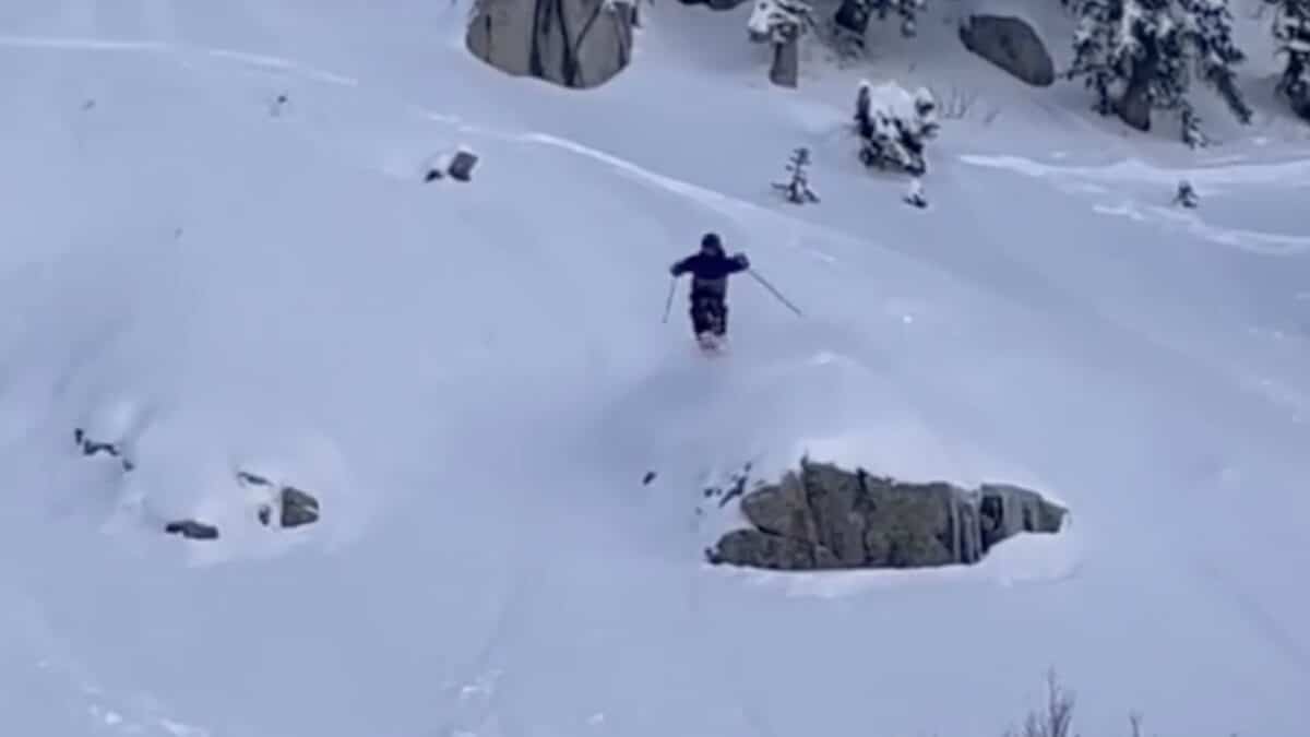 Skier skiing through deep snow on a mountain with rocks and trees around.