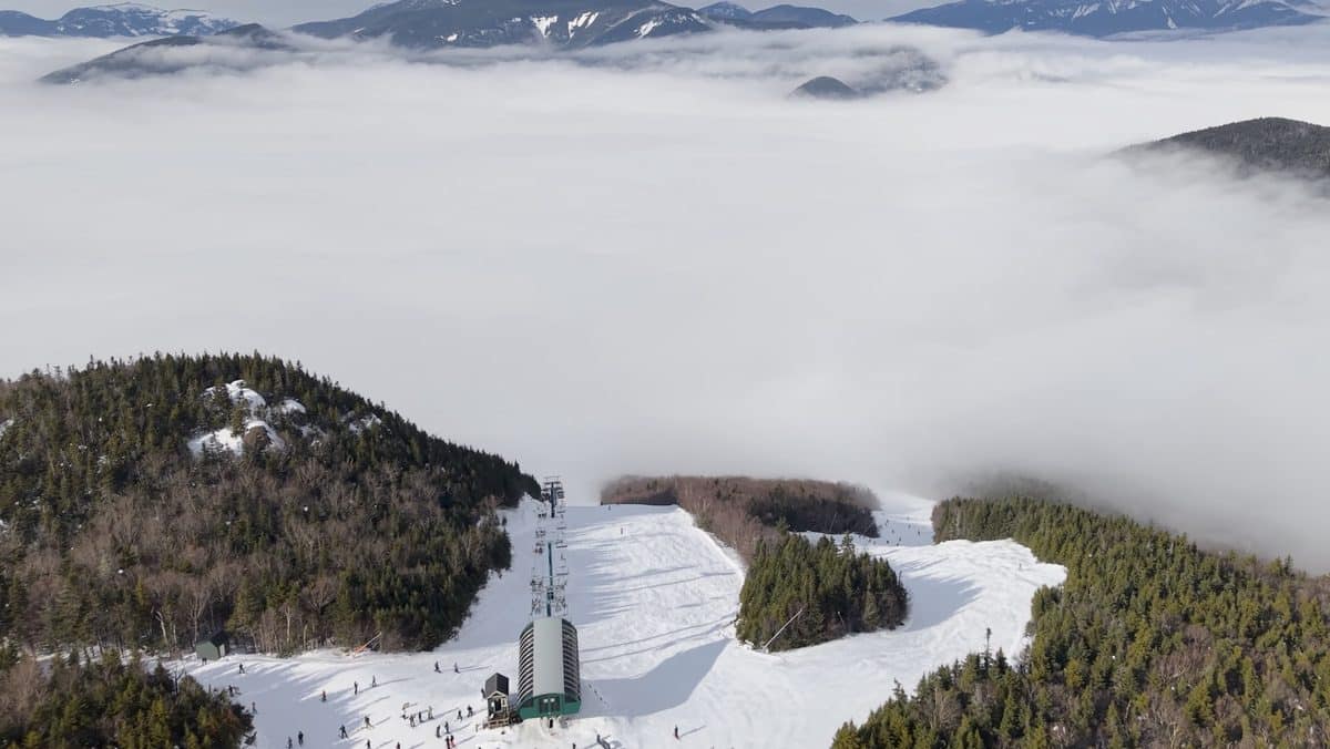 Snowy mountain with ski slopes and forested areas under cloudy sky.