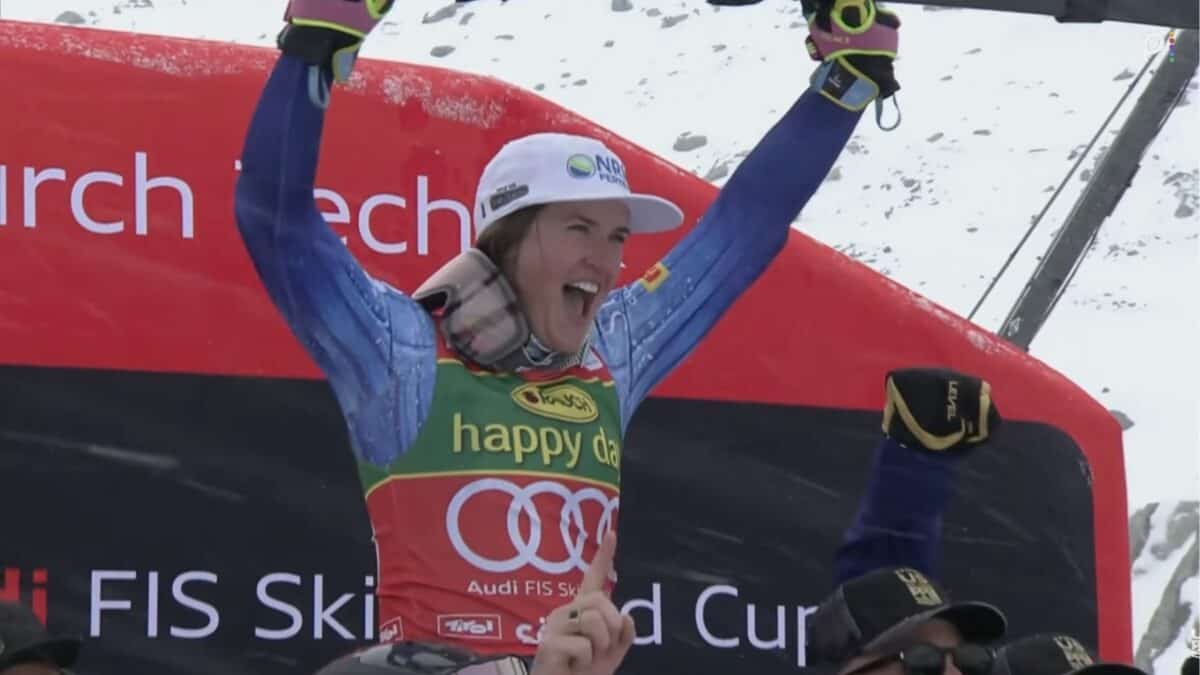 Joyful snowboarder raising arms on winners podium with blue gear and red backdrop.