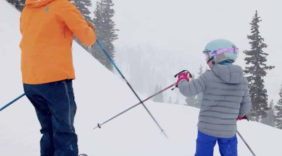 Two skiers in bright jackets on a snowy slope with trees in the background.
