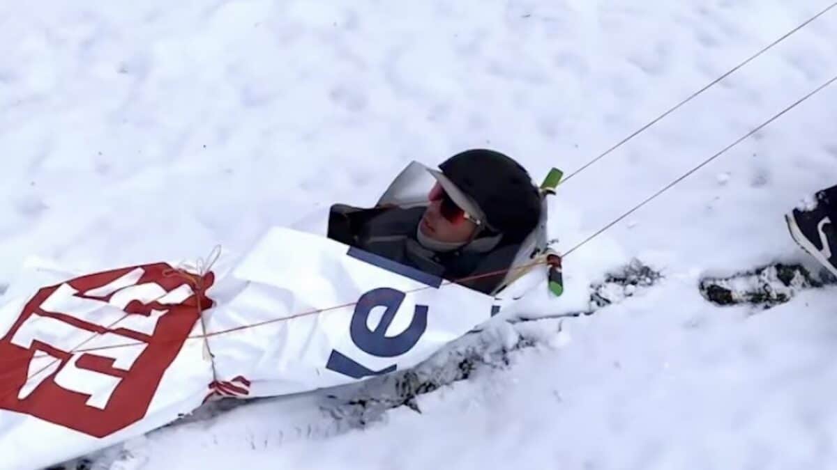 Person in ski gear partially buried in snow with a banner and rescue gear nearby.