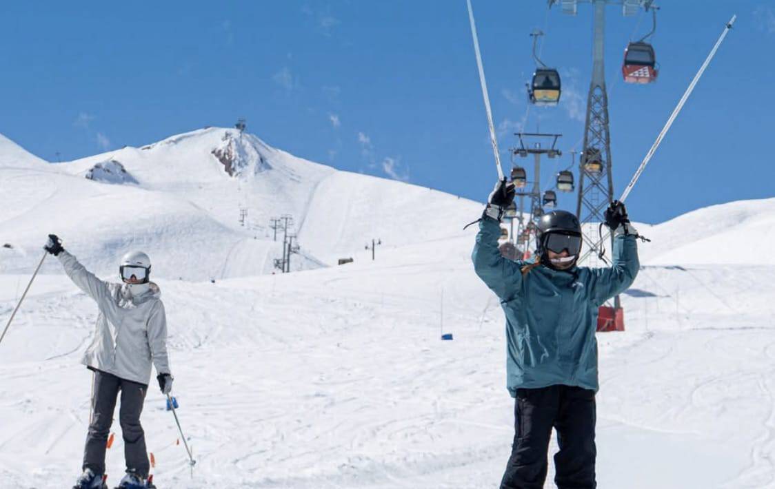 Two skiers on a snowy mountain with a chairlift and blue sky.