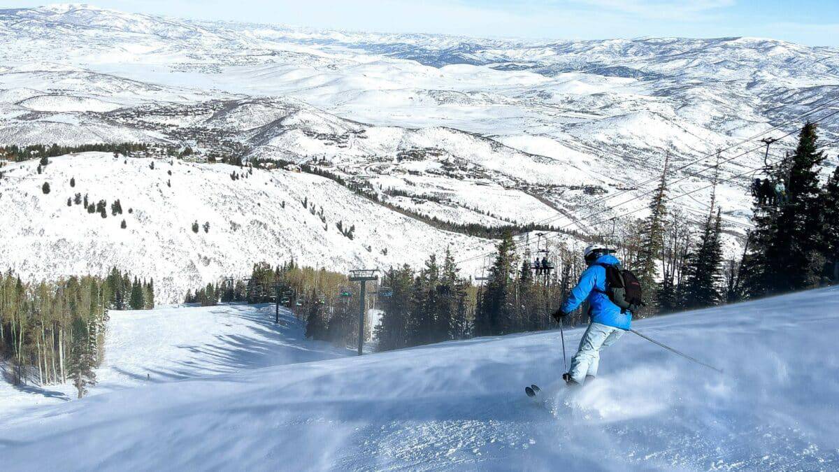 Skier in blue jacket skiing down snowy mountain slope with scenic mountain views.