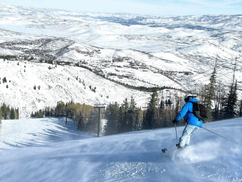 Skier in blue jacket skiing down snowy mountain slope with scenic mountain views.